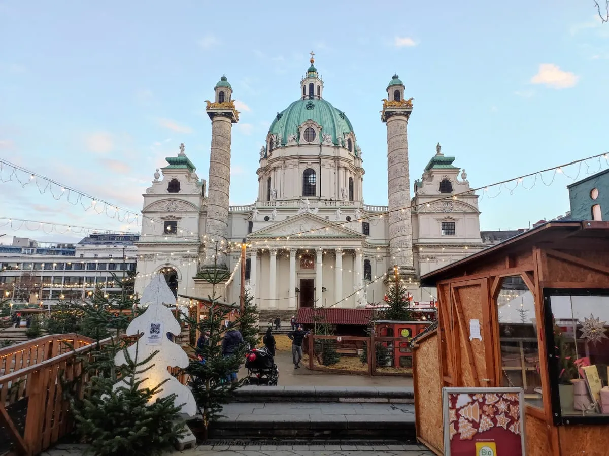 Market huts and string lights in front of the green dome and columns of St. Charles Church at the Art Advent market on Karlsplatz