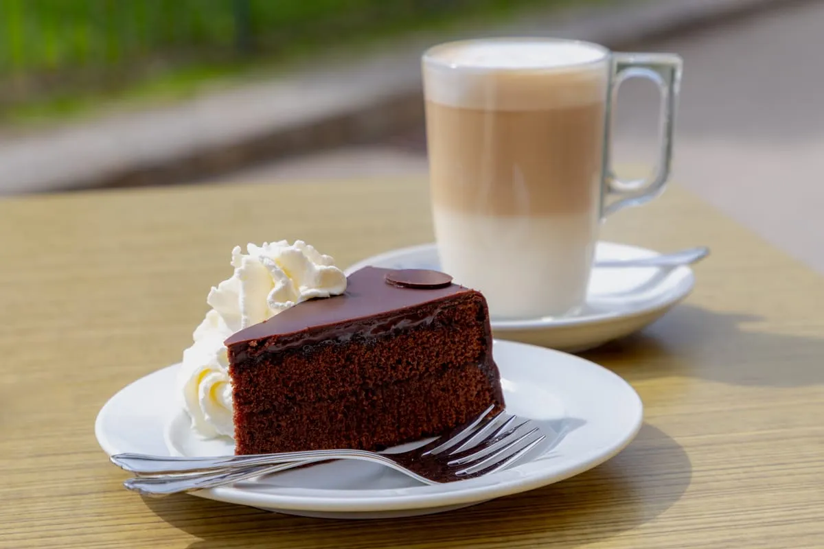 A slice of Sachertorte with whipped cream next to a glass of latte macchiato on a café table