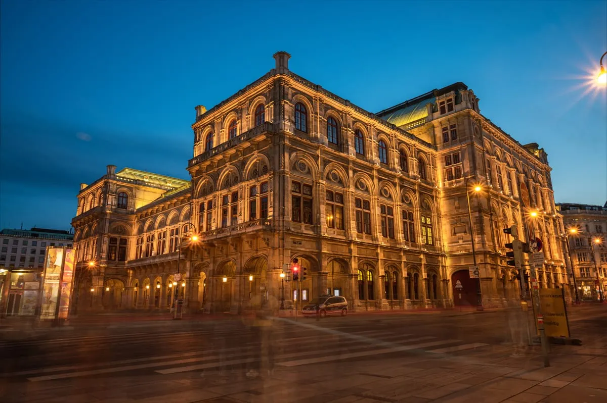 The Vienna State Opera house floodlit at dusk, with light trails from passing traffic on the Ring